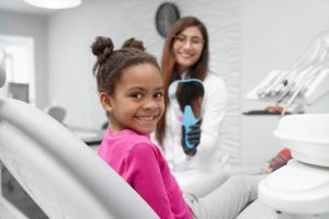 Little african girl sitting on dentist chair looking at camera and smiling while female dentist keeping mirror at backround. Child visiting dentist and enjoying smile after dental procedures.